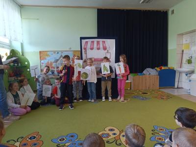 Uczniowie stoją i trzymają w rękach ilustracje z warzywami.