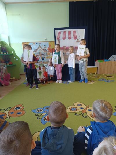 Uczniowie stoją i trzymają w rękach ilustracje z warzywami.