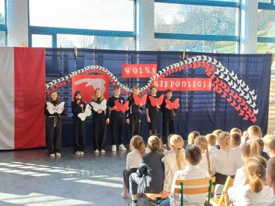 Uczniowie na czarno ubrani recytują i trzymają w dłoniach białe i czerwone motyle.