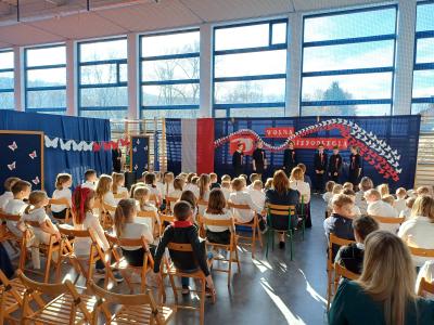 Uczniowie ubrani na czarno recytują, a widownia siedzi na krzesłach.