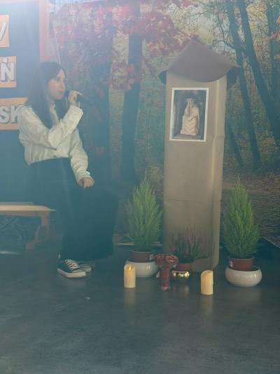 Uczennica siedzi na drewnianej ławeczce i mówi wiersz w kierunku kapliczki z Panem Jezusem Frasobliwym.