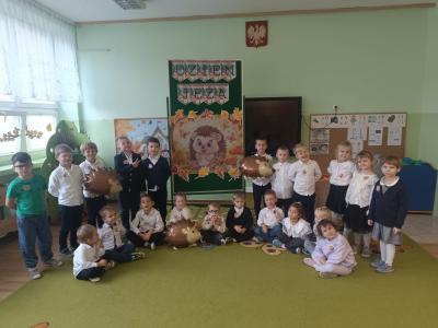 Dzieci stoją i siedzą w grupie, na dywanie po obu stronach obrazka z jeżem i napisem: 