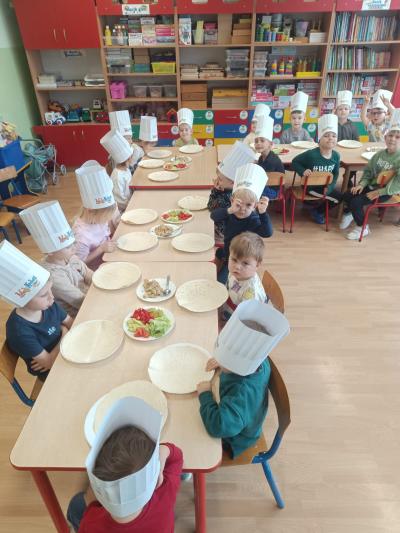 Dzieci w czapkach kucharskich siedzą przy stolikach. Na stolikach znajdują sie nakrycia dla dzieci oraz talerze z warzywami i kurczakiem.