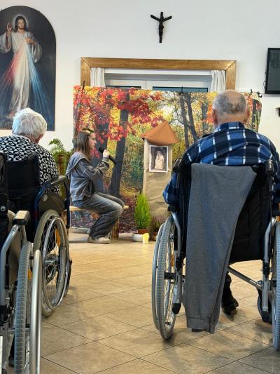 Uczennica siedzi na ławeczce i mówi swoją kwestię do mikrofonu, a osoby chore i starsze słuchają.