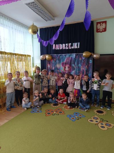 Dzieci stoją i siedzą na tle andrzejkowej dekoracji.