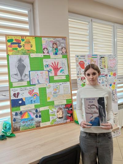 Uczennica trzyma w rękach dyplom. Stoi obok swojej pracy konkursowej.