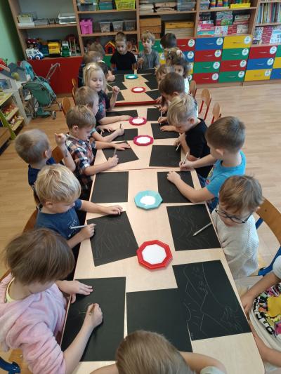 Przedszkolaki siedzą przy stolikach. Na stolikach leżą czarne kartki i sól na talerzykach.