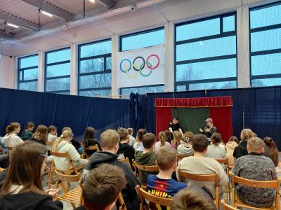Uczniowie siedzą na krzesłach i oglądają spektakl. Na scenie siedzi dwóch aktorów przy ławce szkolnej.