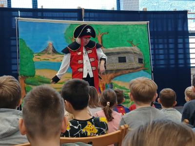 Uczniowie siedzą na krzesłach i oglądają spektakl. Na scenie stoi aktor przebrany za pirata.