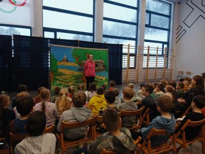 Uczniowie siedzą na krzesłach i oglądają spektakl. Na scenie stoi aktor.