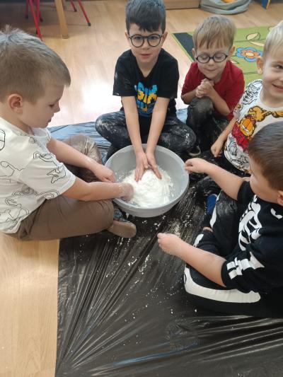 Przedszkolaki siedzą w grupkach na podłodze, a w miskach znajduje się piankolina.