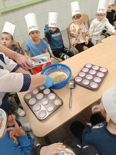 Dzieci przyglądają się, jak kucharka nalewa ciasto do foremek.