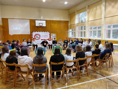 Uczniowie siedzą w kole na krzesłach, na sali gimnastycznej. Niektórzy mają na głowach kolorowe obręcze.