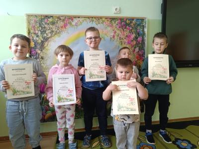 Dzieci trzymają dyplomy, a przed nimi stoją torebki z prezentami.