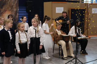 Uczniowie stoją w grupie i śpiewają pieśń. Trzy osoby grają na instrumentach.