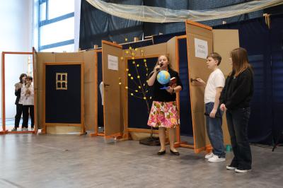 Na scenie uczennica z globusem w ręce i dwóch innych uczniów.