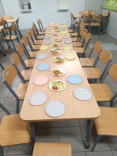 Na stołach na jadalni znajdują się puste talerze dla dzieci oraz talerze z warzywami i kurczakiem.